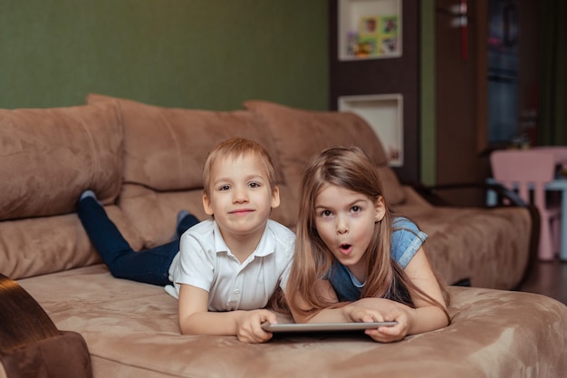 Home distance learning at home. twins brother and sister are studying at home using a tablet. they are happy and laughing while lying on the couch