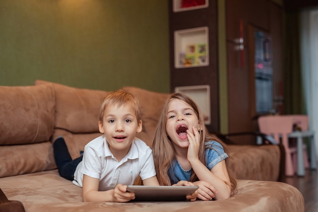 自宅での家庭用遠隔学習。双子の兄と妹は、タブレットを使用して自宅で勉強しています。彼らはソファに横たわっている間幸せで笑っています