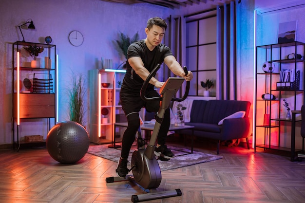 Home fitness allenamento giovane uomo asiatico atleta allenamento su smart cyclette al chiuso