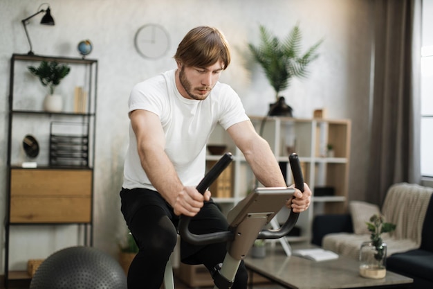 Photo home fitness workout young caucasian man athlete training on smart stationary bike indoors