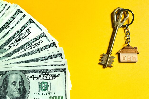 Home key with a keychain on a stack of $ 100 bills on yellow background. Purchase of an apartment, house, real estate, business, mortgage and housing loan from bank, savings, cash, moving. Copy space