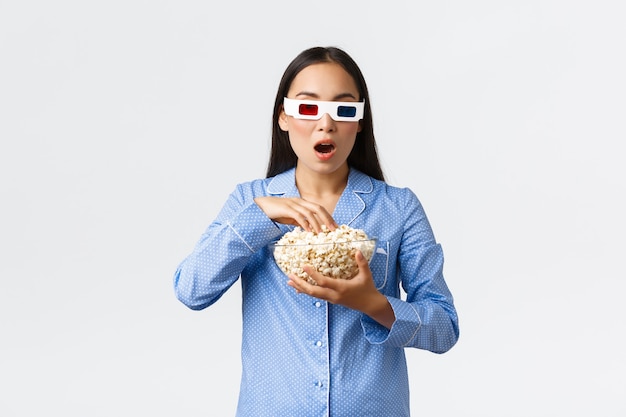 Домашний досуг, ночевка и концепция ночной вечеринки. Пораженная и изумленная молодая азиатская девушка в пижаме и 3D-очках смотрит напряженный момент по телевизору и ест попкорн во время кино.