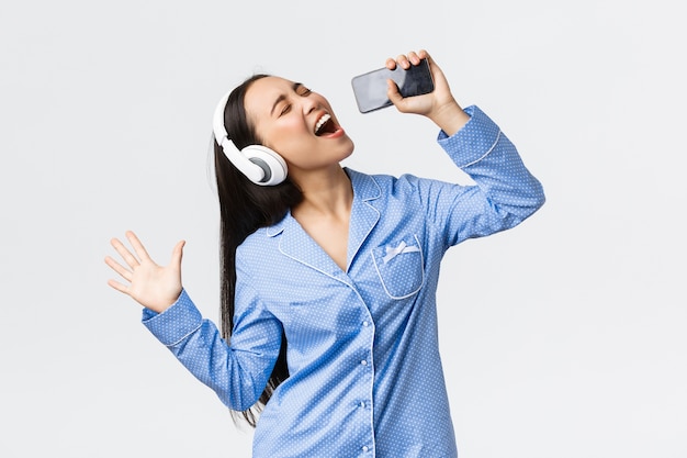 Home leisure, weekends and lifestyle concept. excited and carefree asian girl in pajama, playing karaoke app on smartphone, singing song into mobile phone as wearing headphones, white wall