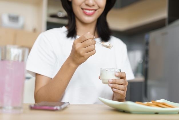 ホームライフスタイルのコンセプトヨーグルトを食べて、家でのんびりしながらスマートフォンを見ている若い女性