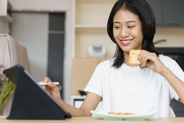 ホームライフスタイルのコンセプトタブレットを使用して、家でのんびりしながらクラッカーを食べる若い女性