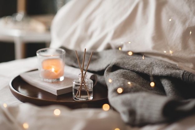 Home liquid fragrance on open paper book and burning scented candle on wooden tray and knit cloth sweater in bed close up Cozy atmosphere