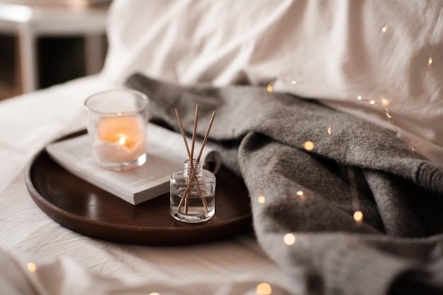 Home liquid fragrance on open paper book and burning scented\
candle on wooden tray and knit cloth sweater in bed close up cozy\
atmosphere