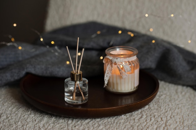 Home liquid perfume in glass bottle and bamboo sticks with\
scented candle with knitted sweater on wooden tray in cozy chair\
over glow christmas lights aromatherapy winter holiday season