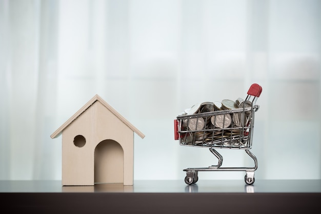 Home and money in the shopping basket on wooden table transparent curtain background. 