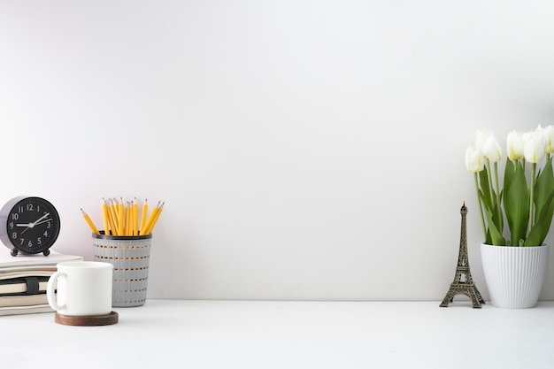 Foto scrivania per l'home office con sveglia libri tazza di caffè portamatite e vaso di fiori su tavolo bianco spazio di copia