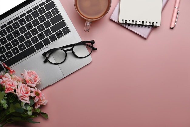 Home office desk with blank notebook laptop computer and pink roses bouquet on pink background Flat lay