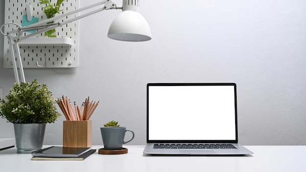 Home office desk with computer laptop, coffee cup, digital tablet, house plant and pencils holder on white table.