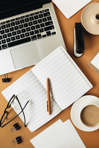 Home office desk workspace with laptop, coffee cup, blank sheet notebook, glasses, pen, green plant branch on ginger