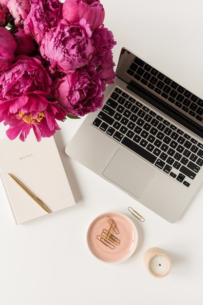 Home office desk workspace with laptop, pink peony flowers bouquet and notebook on white. Flat lay, top view