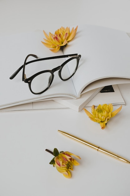 Home office desk workspace with notebook, magazines, glasses, flowers on white table.