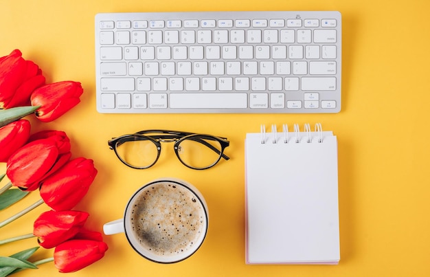 Foto home office sul posto di lavoro tazza di caffè tastiera notebook vuota fiori di tulipano su sfondo giallo disposizione piatta vista dall'alto copia spazio lavoro da casa