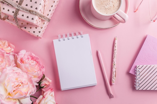 Home office workspace - cup of coffee and fresh rose flowers on pink background. Top view with place for text. Flat lay