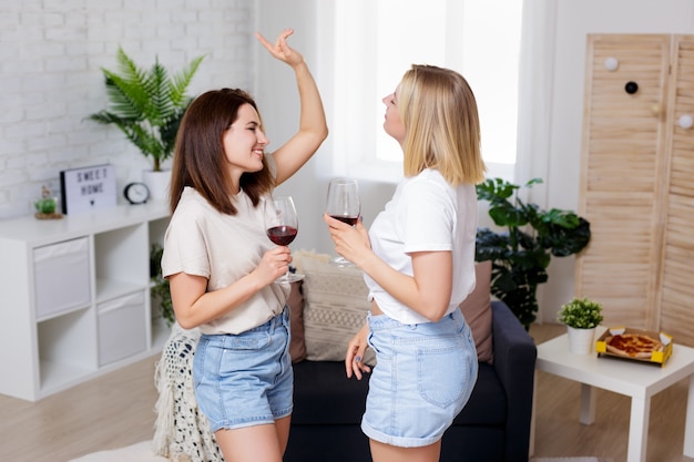 Home party - two beautiful girls having fun together, drinking wine and dancing in cozy living room at home