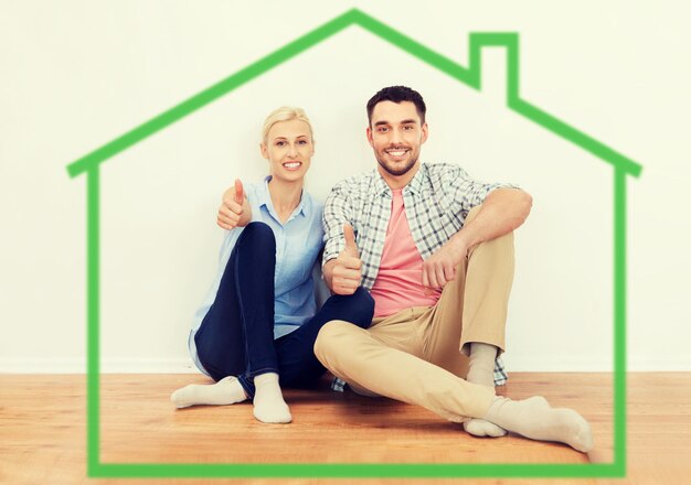home, people, accommodation, moving and real estate concept - happy couple sitting on floor and showing thumbs up at new place