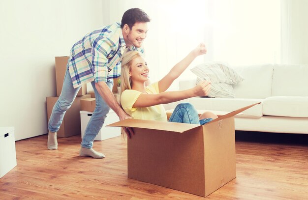 Photo home, people, moving and real estate concept - happy couple having fun and riding in cardboard boxes at new home