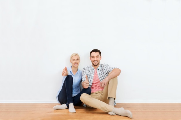 home, people, repair, moving and real estate concept - happy couple sitting on floor and showing thumbs up at new place