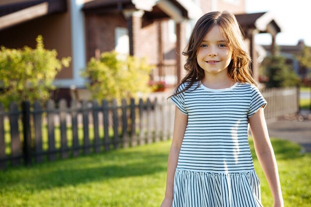 At home. The portrait of a charming petite girl in a striped dress standing near her house and smiling at the camera