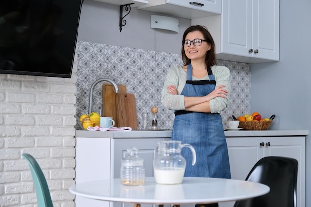 Home portrait of mature woman housewife in apron in kitchen, smiling happy female with folded hands