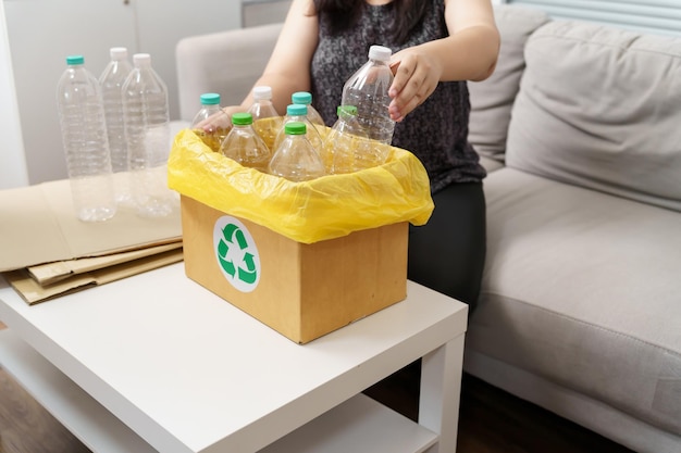 Женщина выбрасывает пустую пластиковую бутылку в контейнер для переработки с желтыми мешками для мусора дома