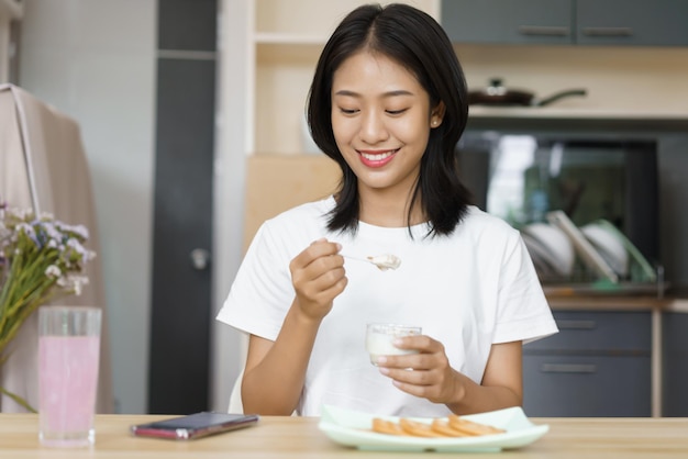 家のリラクゼーションの概念ヨーグルトを食べて、家でのんびりしながらスマートフォンを見ている若い女性