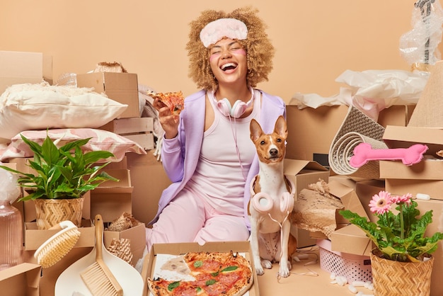 Home relocation concept positive young female tenant surrounded
with belongings moves in new property poses with favorite pet eats
tasty pizza dressed in casual clothes moving day and mortgage