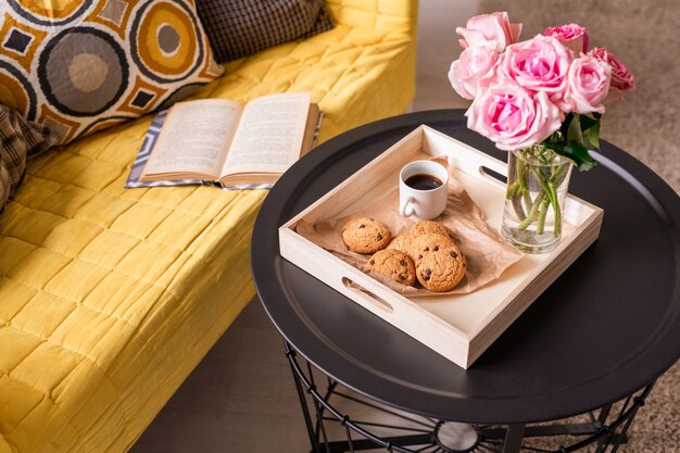 Home still life with bunch of pink roses, homemade snack and drink on small table and open book and cushions on yellow couch