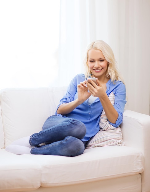 Concetto di casa, tecnologia e internet - donna sorridente con smartphone seduto sul divano di casa