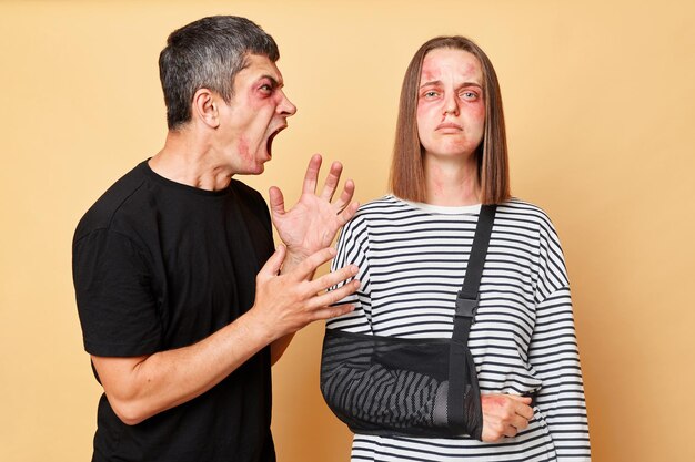 Home violence Relationship arguingg Aggressive man screaming with hate and anger beating his woman wife with bruise and bandage isolated over beige background