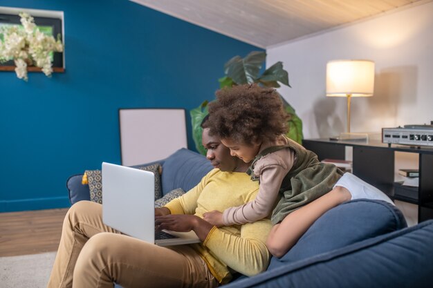 Home weekdays. Concentrated african american dad looking at laptop and bored little daughter hugging shoulders sitting on sofa in modern room