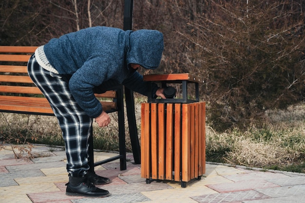 homeless elderly old Caucasian man rummages for food and garbage in trash can in a park in autumn