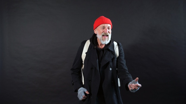 Бездомный, пенсионер, старик с седой бородой в пальто и красной шляпе на изолированном темном фоне