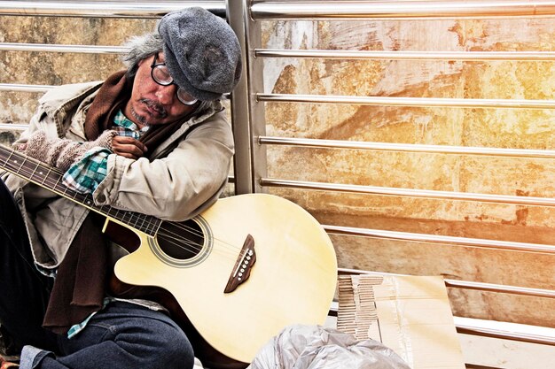 Foto uomo senzatetto seduto con la chitarra contro la ringhiera