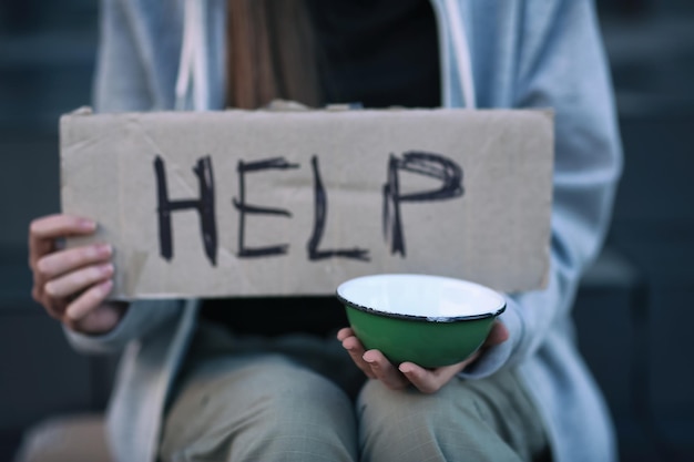 Homeless poor woman holding empty bowl and piece of cardboard with word HELP outdoors closeup