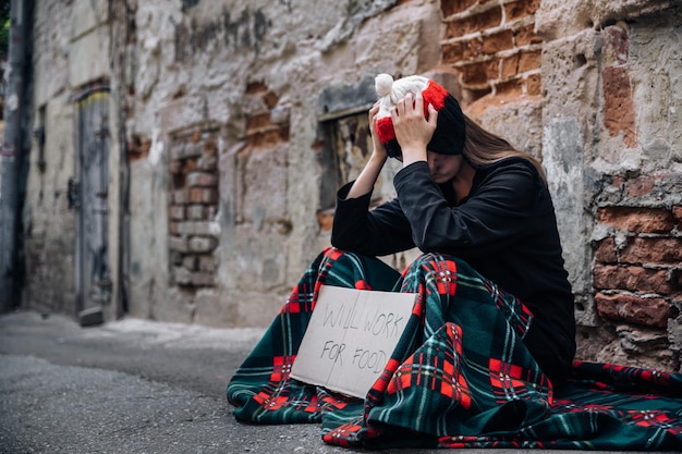 A homeless woman sits on the street in the shade of a house and asks for help and money Problems of large modern cities Grasp your head with your hands Poverty drugs