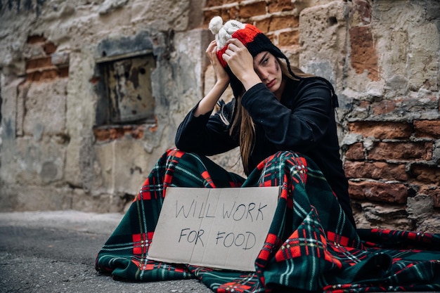 A homeless woman sits on the street in the shade of a house and