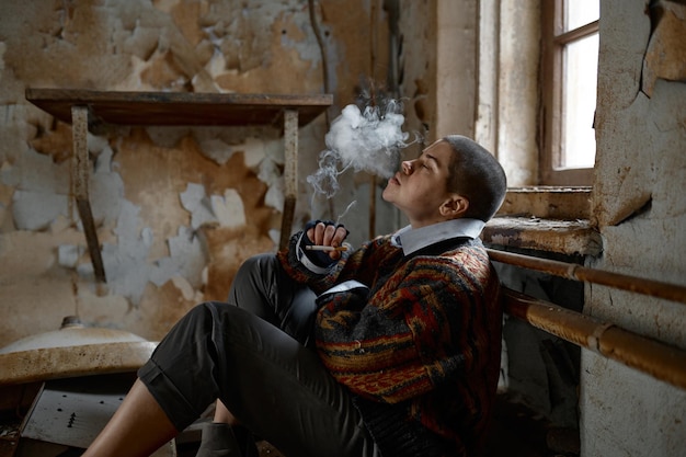 Homeless woman smoking sitting on room floor of abandoned house and looking at camera