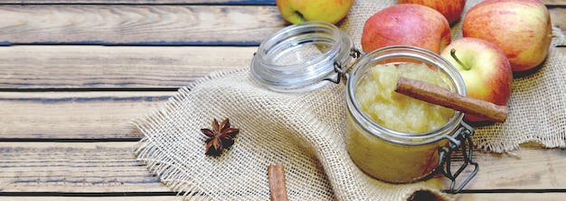 Homemade apple sauce in a glass jar with red apples and stick of cinnamon on a wooden table