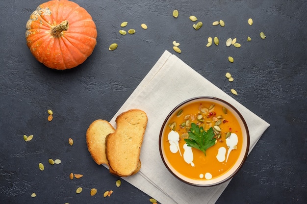 Homemade autumn pumpkin cream soup with cream, croutons, seeds and basil on linen napkin on light wooden background.