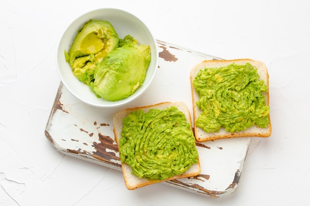 Homemade avocado toasts and smashed fresh ripe avocados in bowl on wooden rustic cutting board on white background. Avocado smash sandwiches, healthy snack good for diet, clean eating, top view