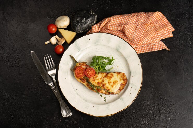 Фотография домашние запеченные баклажаны с сыром и помидорами. горячее блюдо из запеченных овощей с зеленым салатом из рукколы и тимьяна на черном столе. вид сверху.