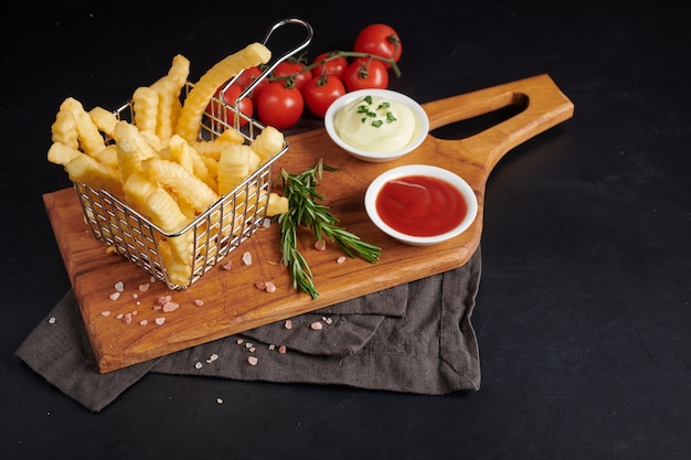 Homemade baked potato fries with mayonnaise, tomato sauce and rosemary on wooden board. tasty french fries on cutting board, in brown paper bag on black stone table background, unhealthy food.