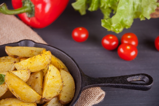 Фото Домашние запеченные картофельные дольки с травами на сковороде черного железа с овощами на заднем плане.