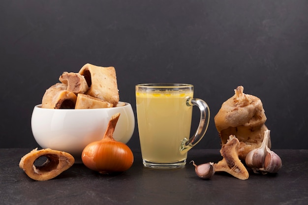 Homemade boiled beef bone broth in transparent cup on dark  background with meat and vegetables. B