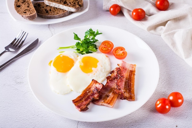 Homemade breakfast fried bacon and eggs tomatoes and greens on a plate on the table