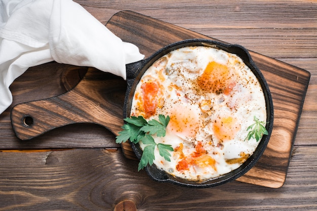 Homemade breakfast of shakshuka fried eggs with tomatoes and herbs in a pan on a wooden table